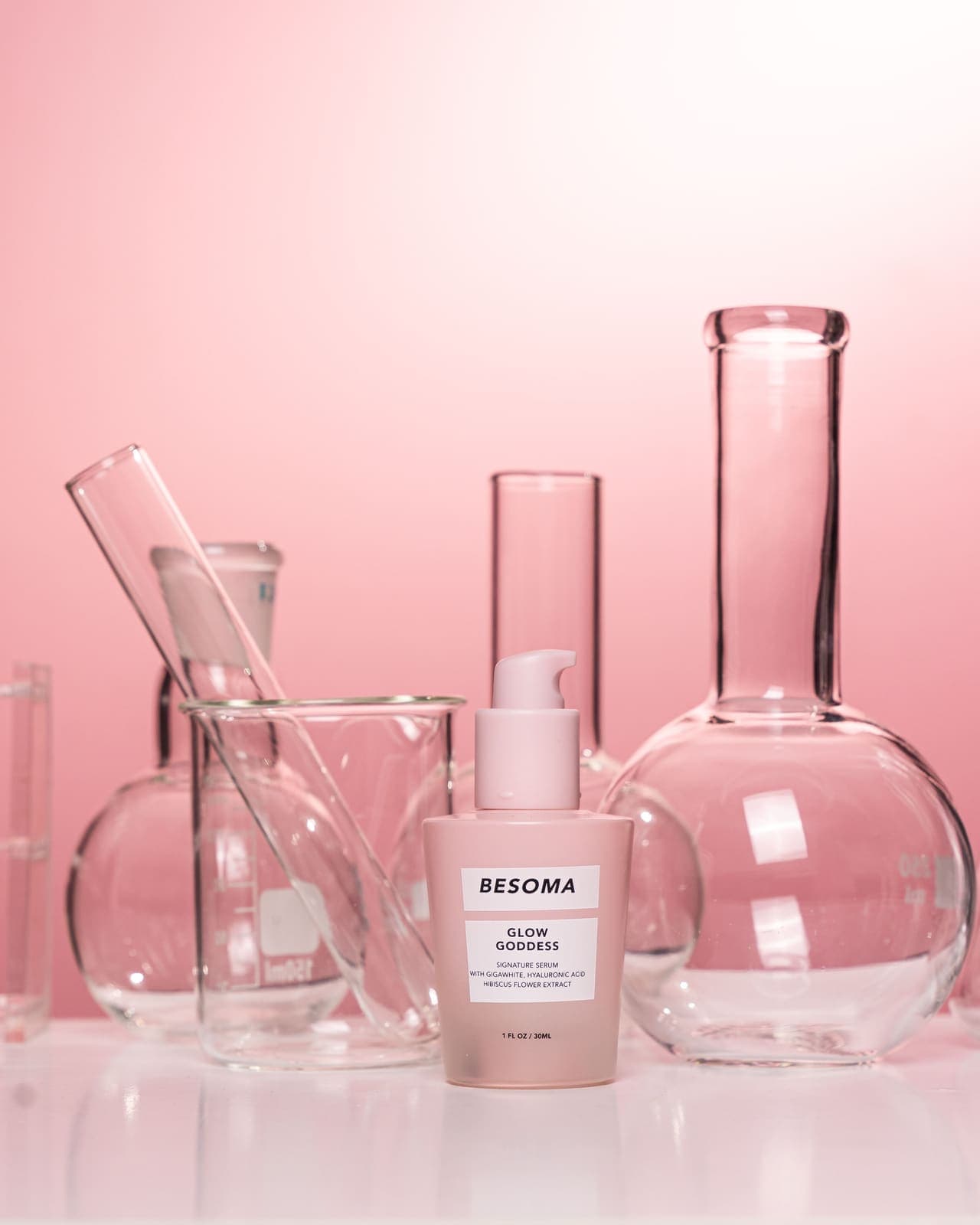 A skincare bottle styled with scientific glassware against a pink background.