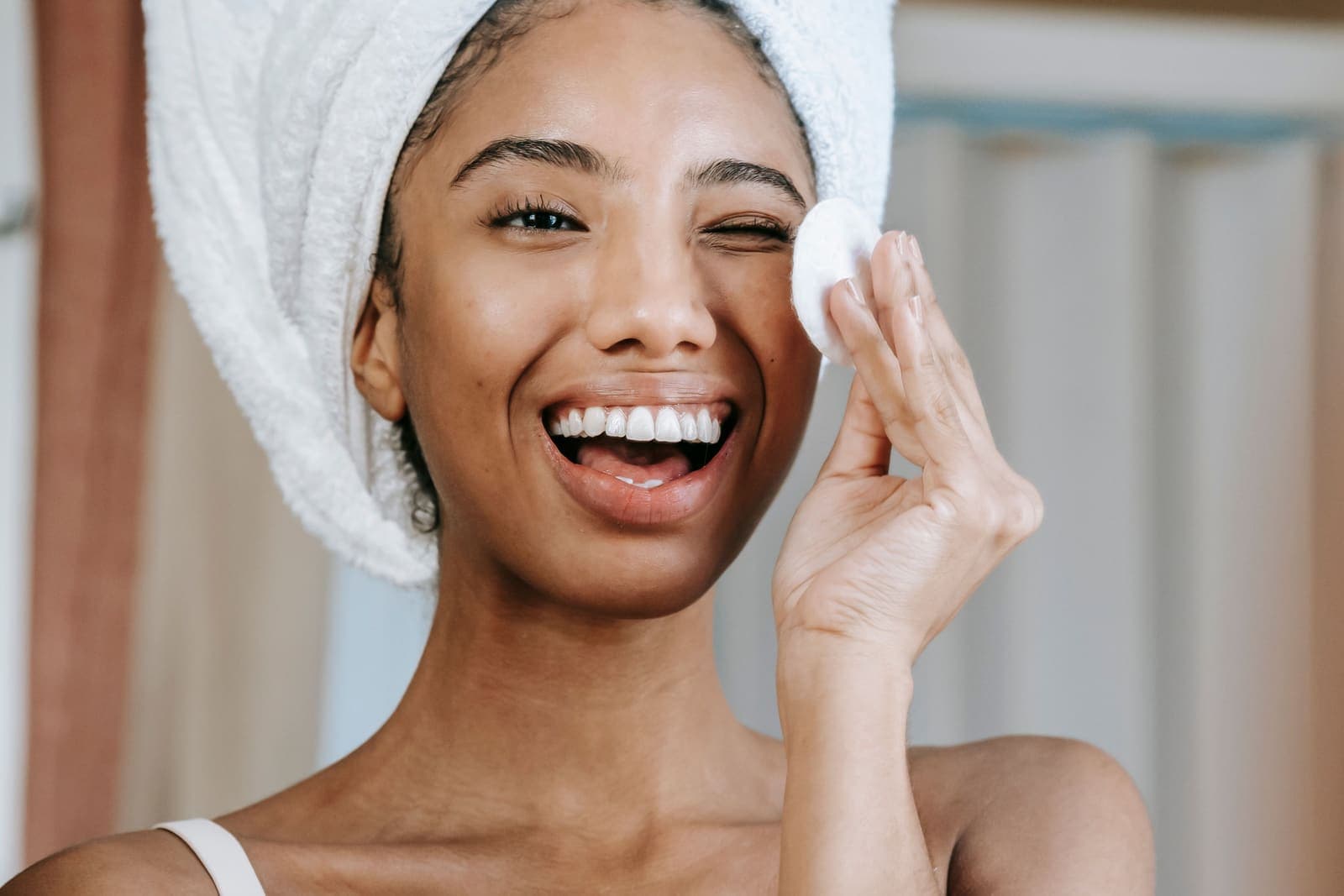 A woman smiling while cleansing her face with a cotton pad in the morning.